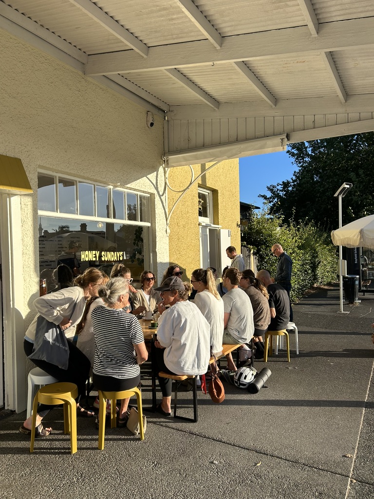 A long table of people chatting over coffee outside Honey Sundays café after a Stretchy Yoga session, yoga mats at their feet
